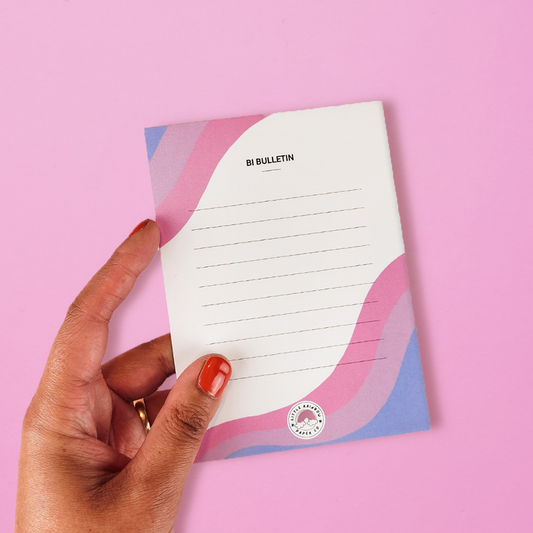A hand holds a LGTBQ notepad with the bi pride flag colors printed on the front. Text reads bi bulletin and has lines for writing. A cute bisexual notepad