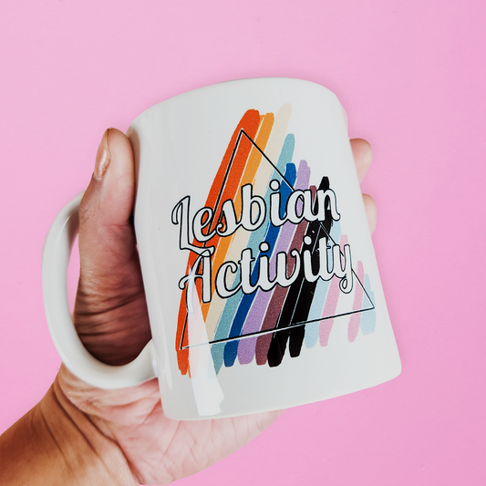 Hand holding LGBTQ mug that reads Lesbian Activity over progress pride colors. Cute lesbian mug on pink background