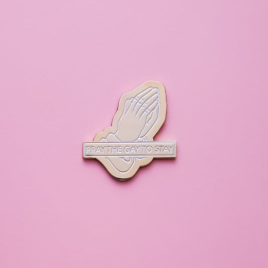 A gold praying hands shaped enamel pin set on a pink background. This pride pin reads 'Pray The Gay To Stay' in white enamel set in a polished gold metal. Pray the Gay to Stay Pin - LGBTQ Pins For Sale | Little Rainbow Paper Co